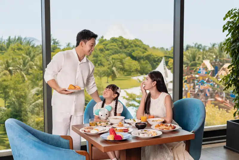 Gia đình ba người ăn sáng bên cửa kính nhà hàng The Blue, bên ngoài là núi Phú Sĩ thu nhỏ và tàu cướp biển công viên nước, Mikazuki Đà Nẵng