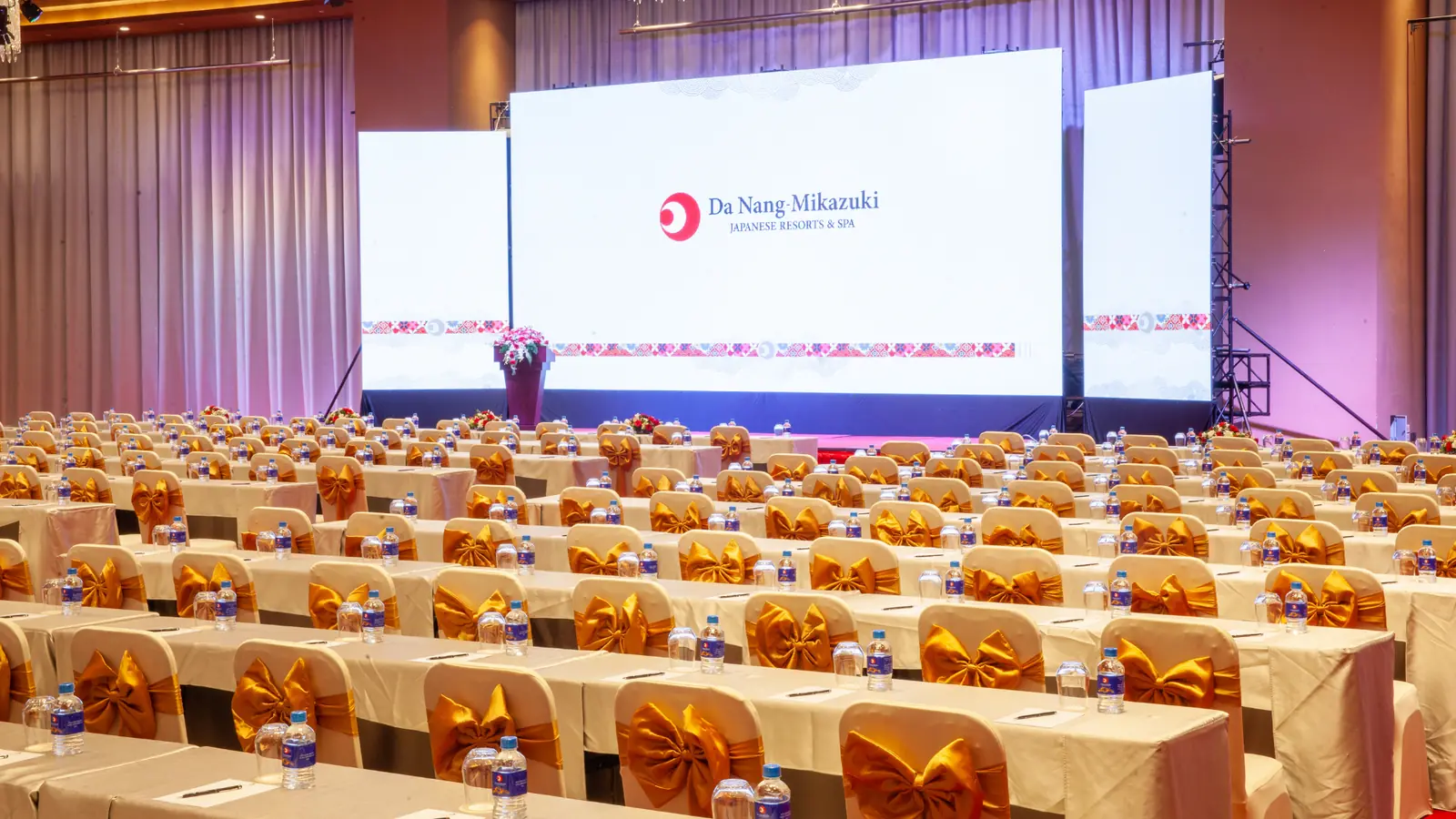 Nippon Ballroom Mikazuki Đà Nẵng bày bàn kiểu lớp học phủ khăn trắng, nơ vàng, hướng về ba màn LED trên sân khấu