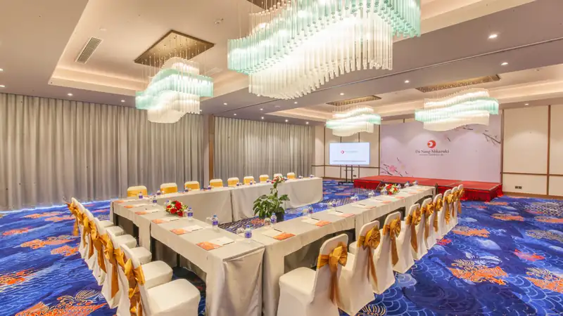 Phòng Sakura Mikazuki Đà Nẵng bày bàn chữ U hướng về sân khấu thảm đỏ, đèn chùm pha lê, thảm xanh hoa văn sóng