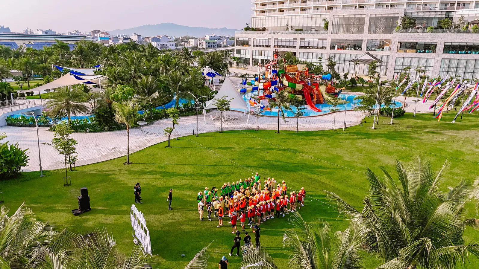 Đoàn team building xếp hàng trên bãi cỏ, phía sau là khu máng trượt ngoài trời và tòa khách sạn Mikazuki Đà Nẵng, nhìn từ trên cao
