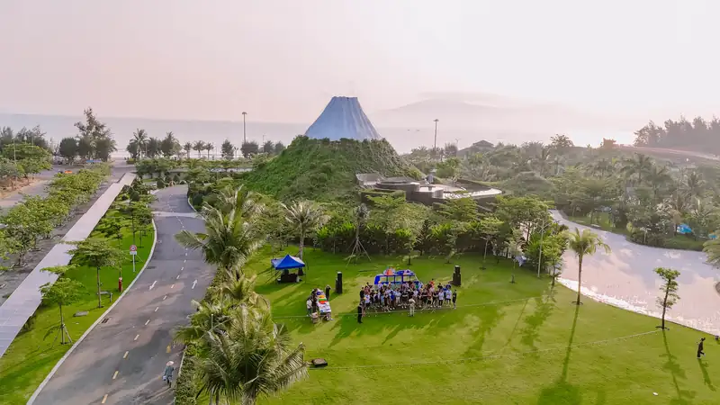 Bãi cỏ rộng có lều xanh, loa và một nhóm khách team building, phía sau là núi Phú Sĩ thu nhỏ và biển, Mikazuki Đà Nẵng