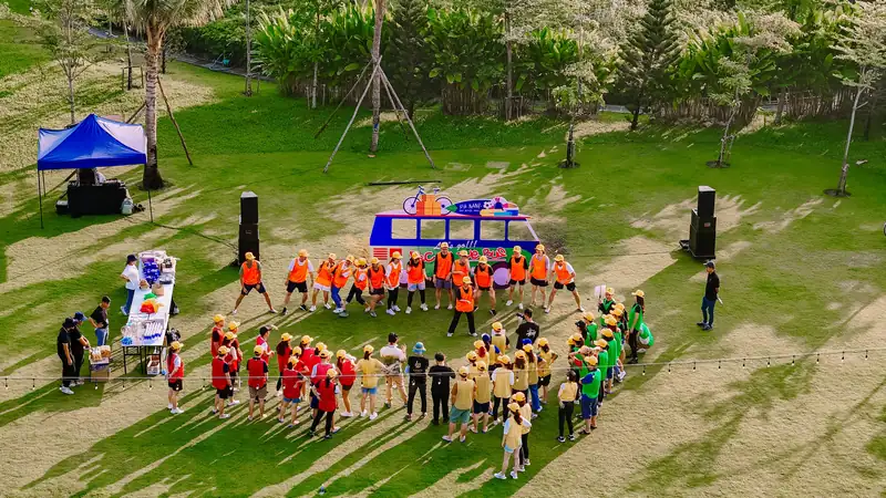 Các đội team building áo cam, đỏ, vàng, xanh đứng thành vòng quanh mô hình xe buýt trên bãi cỏ có loa và lều quản trò