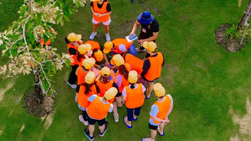 Một đội team building mặc áo yếm cam, đội mũ vàng chụm đầu bàn chiến thuật trên bãi cỏ, nhìn từ trên cao