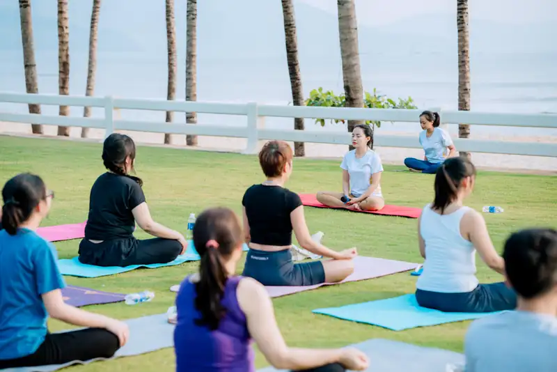 Nhóm khách nữ ngồi thiền trên thảm yoga ở bãi cỏ cạnh bãi biển, hàng dừa và biển phía sau, Mikazuki Đà Nẵng