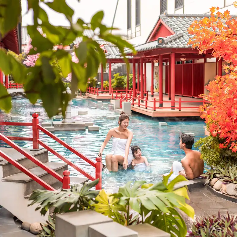 Gia đình ngồi trên bậc đá bể onsen ngoài trời cạnh cầu thang lan can đỏ, Mikazuki Đà Nẵng