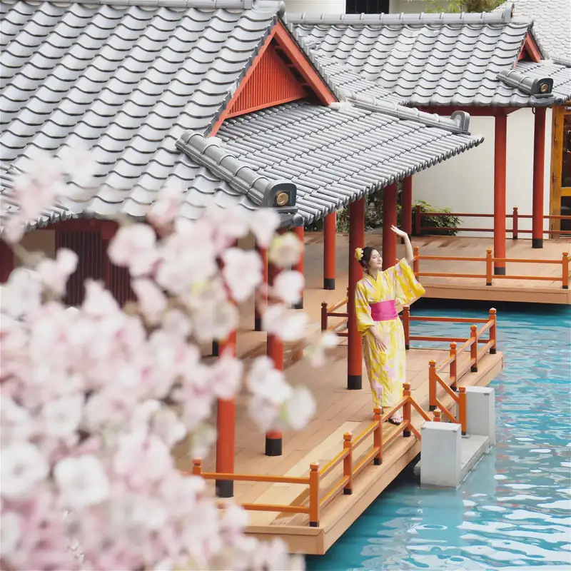 Khách mặc yukata vàng đứng trên hiên gỗ bể onsen dưới mái ngói, hoa anh đào phía trước, Mikazuki Đà Nẵng