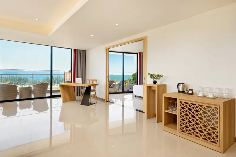 Tủ trà gỗ chạm hoa văn và góc làm việc trong Executive Suite, cửa kính nhìn ra biển Đà Nẵng