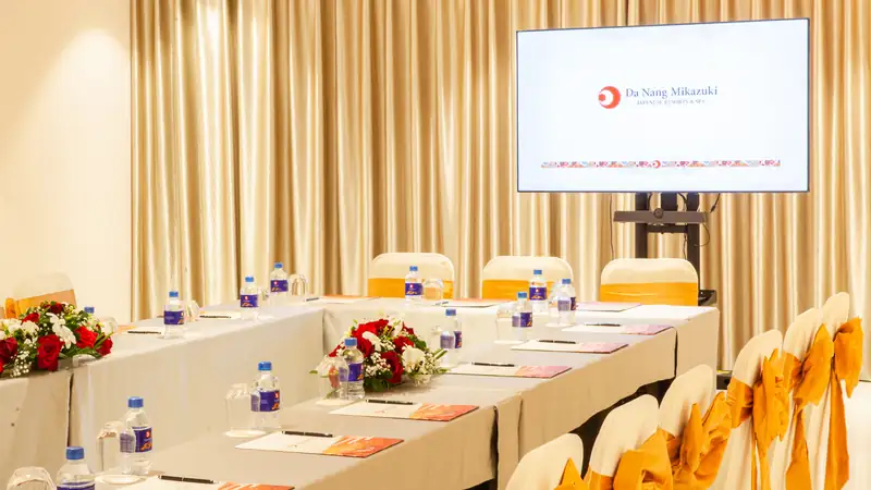 Phòng Hasu bày bàn họp phủ khăn trắng, hoa hồng và nước suối, màn hình hiện logo Da Nang Mikazuki