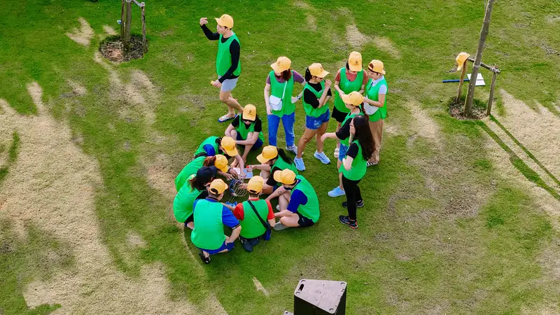 Một đội team building áo xanh, mũ vàng họp nhóm trên bãi cỏ, nhìn từ trên cao